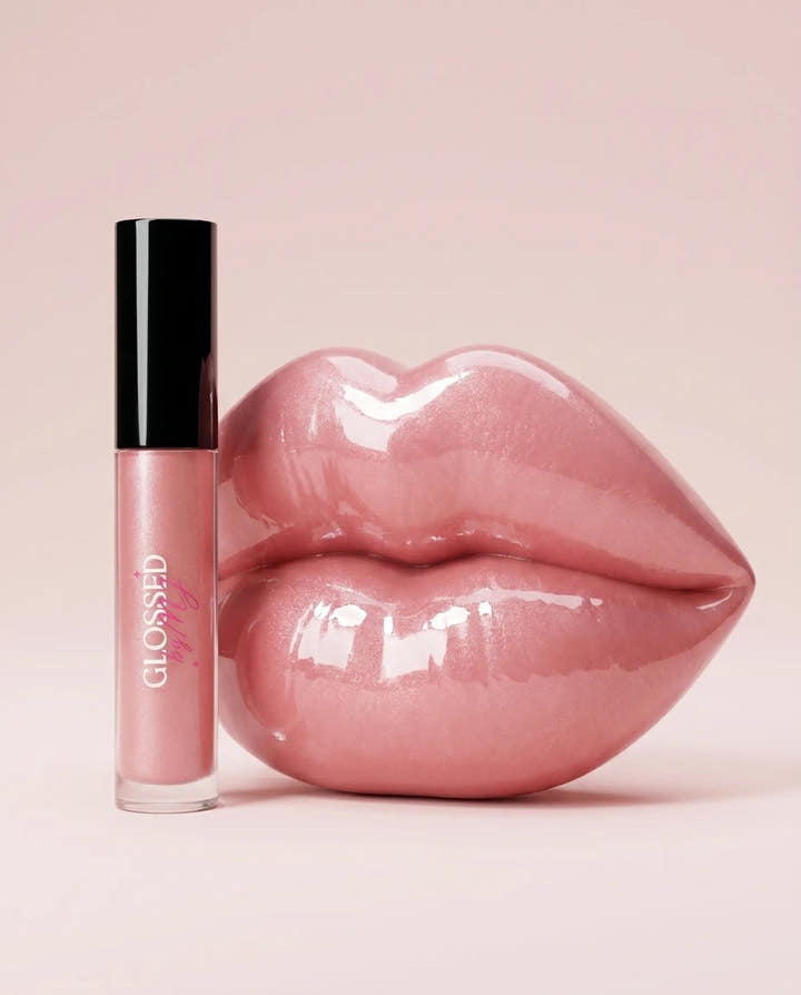A Glossed by MJ lip gloss tube standing beside a glossy sculpted pair of pink lips on a soft blush background.